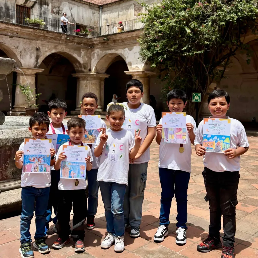 Alumnos de Kiddies Club mostrando sus diplomas y dibujos premiados en un concurso de dibujo y pintura, en un patio colonial de La Antigua Guatemala