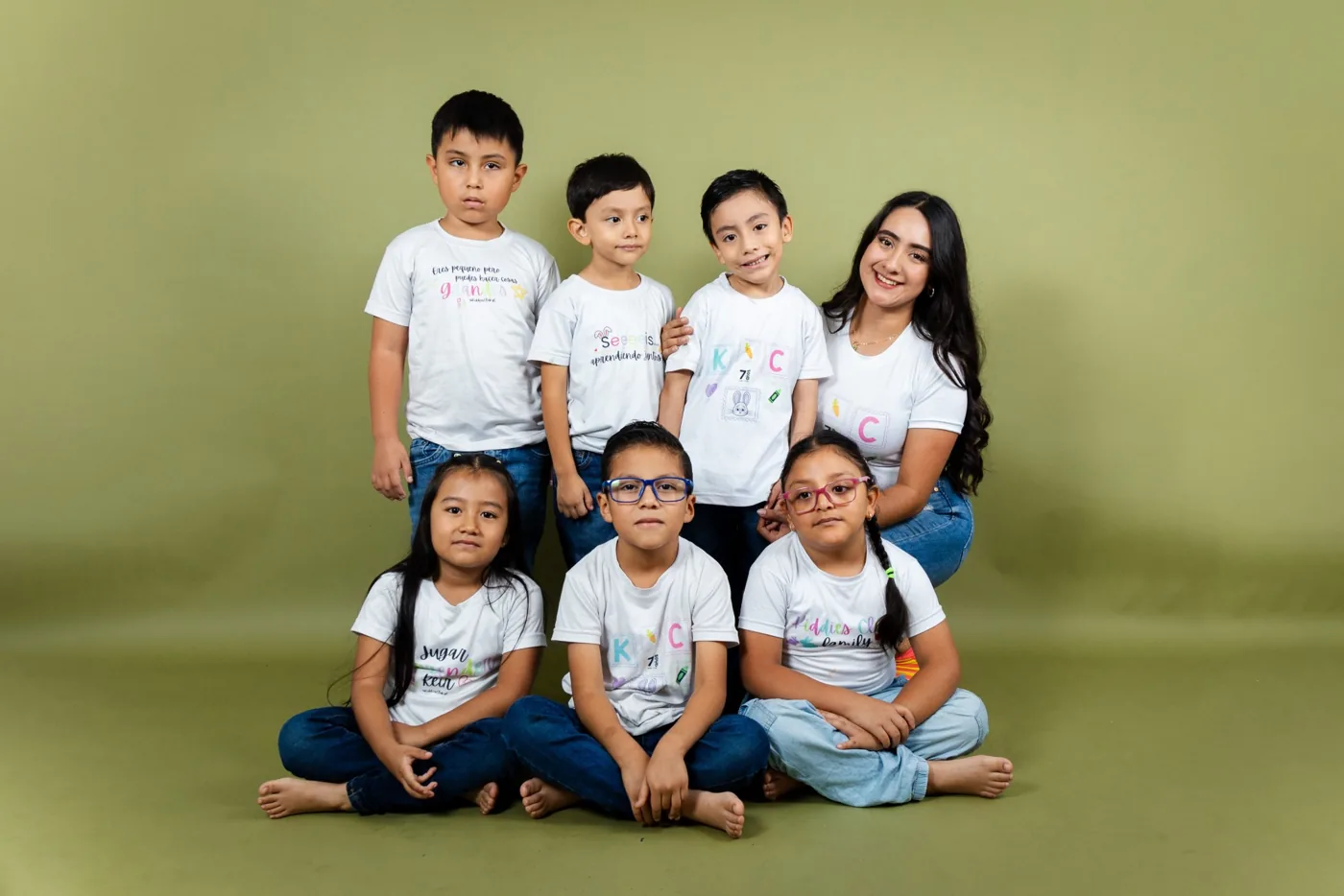 Grupo de alumnos de Kiddies Club con su maestra, sonriendo con sus camisetas de Kiddies en una sesión de fotos